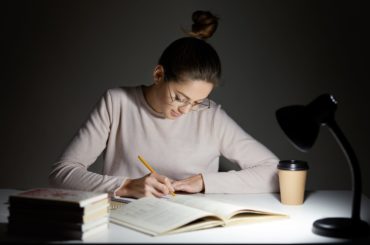 Busy freelancer rewrites infrormation into notepad, prepares article for publication, reads books, writes some notes in organizer, drinks takeaway coffee, sits in darkness, wears round big spectacles