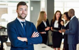 Caucasian businessman leader looking at camera in modern office with multi-ethnic businesspeople working at the background. Teamwork concept. Young man with beard wearing blue suit.