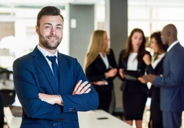Caucasian businessman leader looking at camera in modern office with multi-ethnic businesspeople working at the background. Teamwork concept. Young man with beard wearing blue suit.