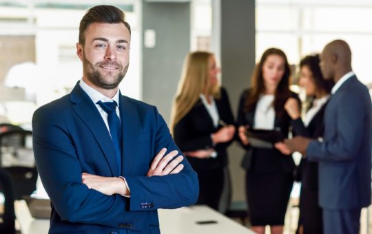 Caucasian businessman leader looking at camera in modern office with multi-ethnic businesspeople working at the background. Teamwork concept. Young man with beard wearing blue suit.